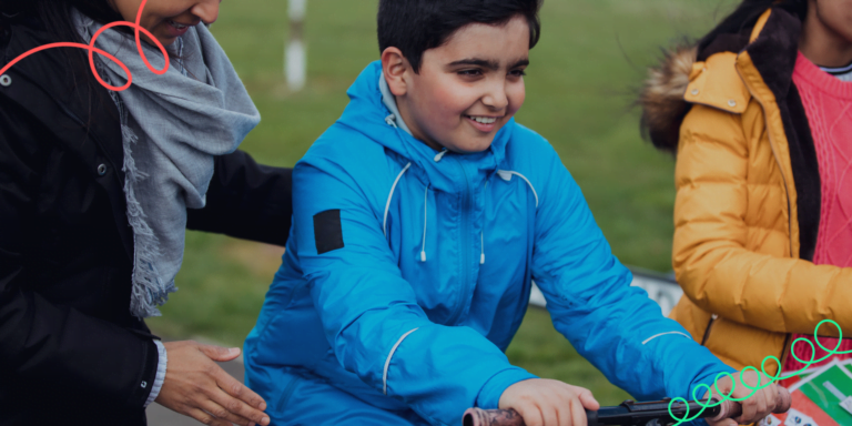 A young boy smiling on a bicycle being supported by an adult